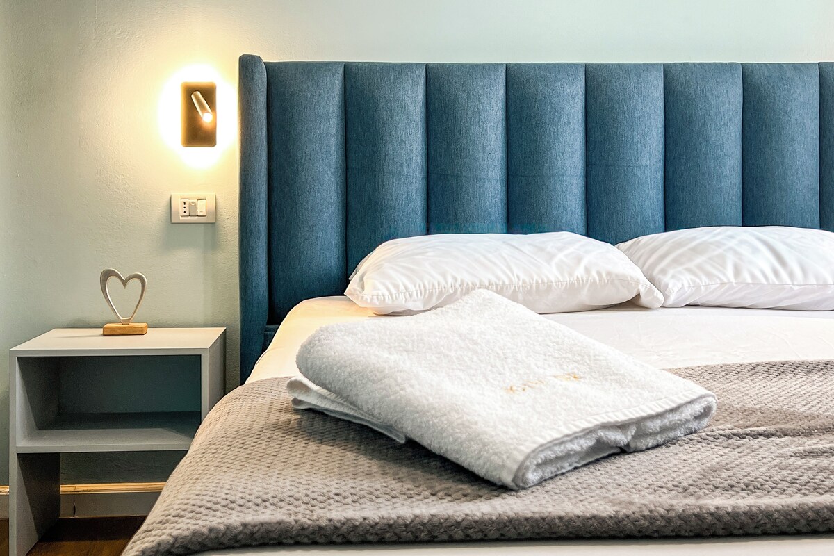 A neatly made bed with white linens and a folded towel, a bedside table with a heart-shaped decoration, and a wall-mounted light.