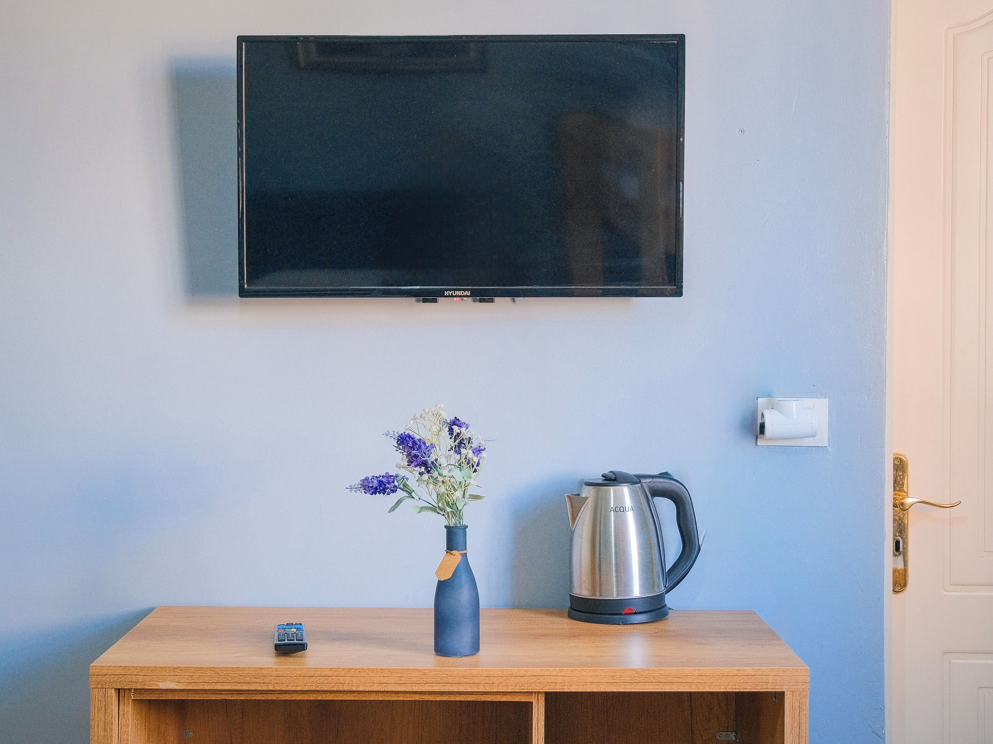 Ein Zimmer mit Holztisch, Blumen in einer Vase, einem Wasserkocher und einem an der Wand montierten Fernseher.