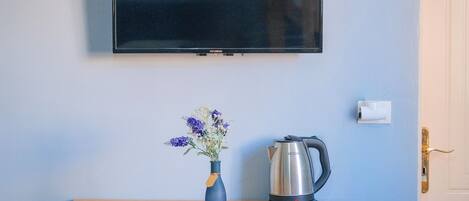 Ein Zimmer mit Holztisch, Blumen in einer Vase, einem Wasserkocher und einem an der Wand montierten Fernseher.