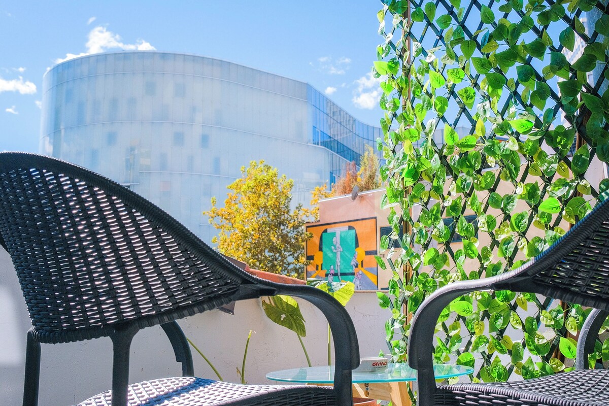 Dos sillas de mimbre con una mesa de vidrio, una planta en maceta y un edificio con un mural al fondo.