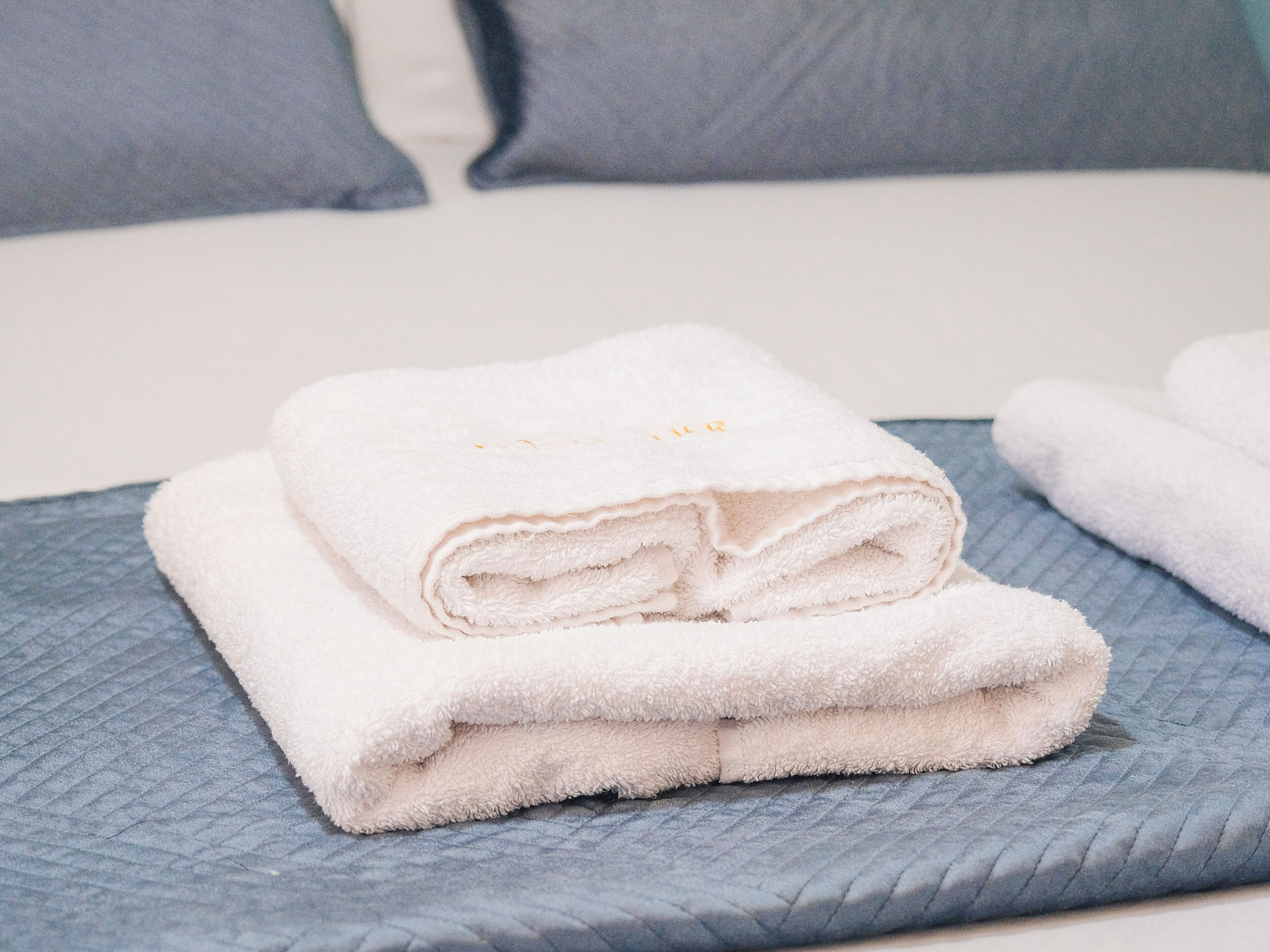 Une serviette blanche, soigneusement pliée, est posée sur un couvre-lit bleu matelassé, avec un oreiller gris en arrière-plan.