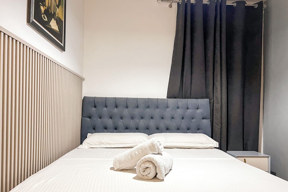 A neatly made bed with white linens and two rolled towels, a dark upholstered headboard, a bedside table, and a framed picture on the wall.