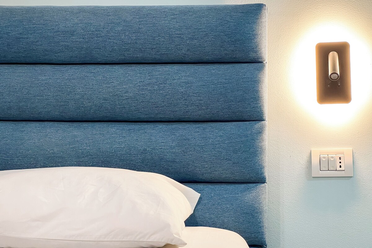 A bed with a blue headboard and a white pillow, a wall-mounted light, and a power outlet.