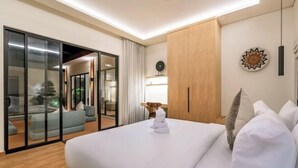A modern hotel room with a large bed, a sliding glass door leading to a balcony, a wooden wardrobe, and a wall-mounted lamp. - Oasis Residences Sanur – Family-Friendly 3-BR Villa with Private Pool & Garden (Bali)