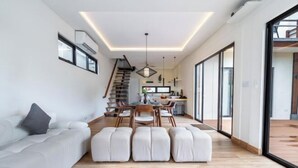 A modern living space with a sofa, ottomans, a dining area, and a staircase. - Oasis Residences Sanur – Family-Friendly 3-BR Villa with Private Pool & Garden (Bali)