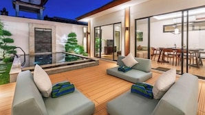 A modern outdoor living area with a wooden deck, two grey sofas with cushions, a glass door leading to a dining area, and a traditional-style structure in the background. - Oasis Residences Sanur – Family-Friendly 3-BR Villa with Private Pool & Garden (Bali)