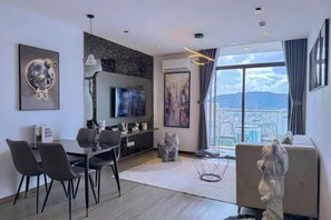 A modern living room with a TV, dining area, and a large window with a city view. - Meo's House - CSJ Seaside Apartment (Vung Tau)