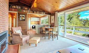 A cozy cabin interior with a fireplace, comfortable seating, a dining area, and a large window offering a view of the outdoors.