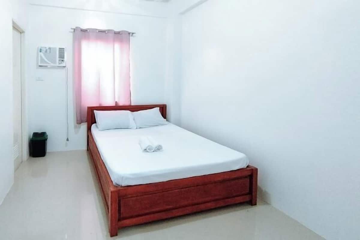 A single-bed room with a wooden frame, white bedding, a small bedside table, a trash bin, and a window with a curtain.