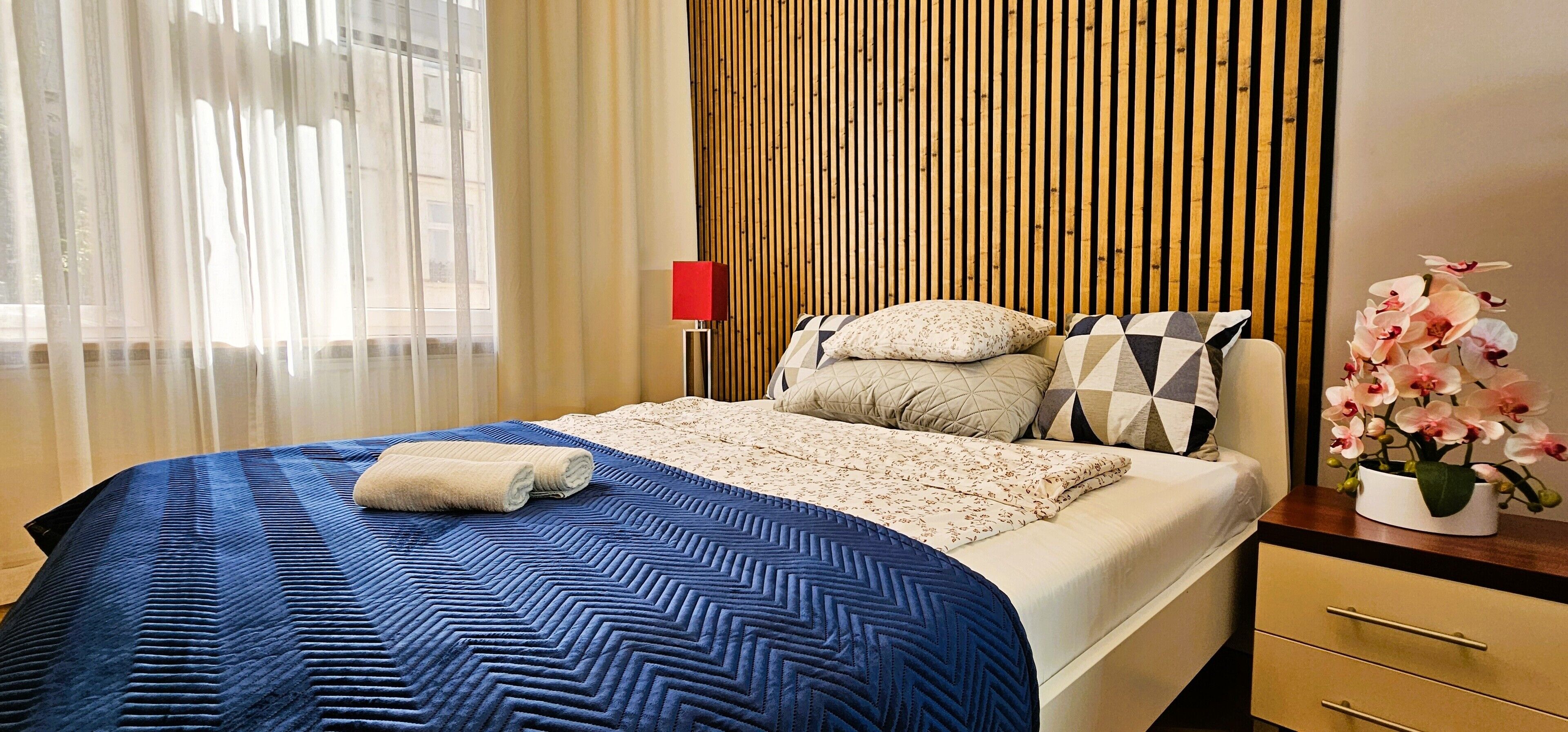 A modern hotel room with a large bed, a wooden slat wall, a red floor lamp, and a vase of flowers.