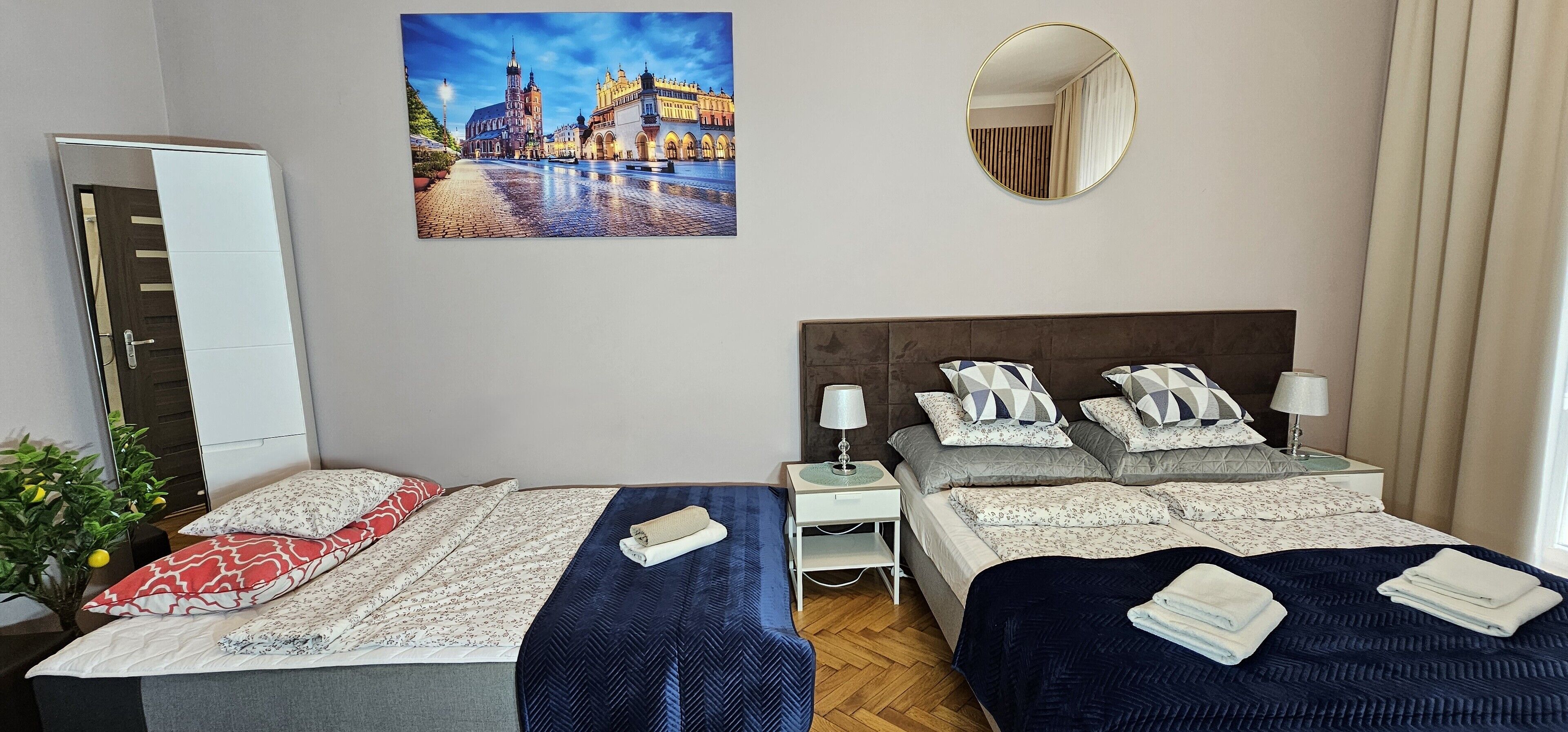 A hotel room with two beds, a painting of a cityscape, a round mirror, and a plant.