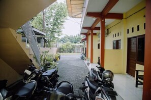 A covered outdoor area with motorcycles parked, a tiled floor, and a yellow building with a door and windows. - RedDoorz @ Hening Homestay (Gunung Sari)