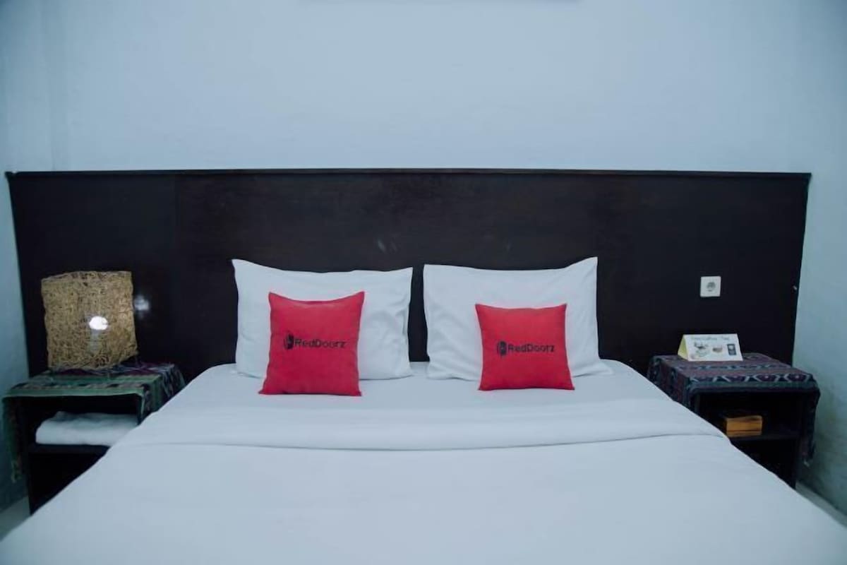 Una cama con sábanas blancas, dos almohadas rojas, un cabecero oscuro y una mesita de noche con una lámpara.