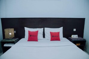 A bed with white sheets, two red pillows, a dark headboard, and a small table with a lamp on the side. - RedDoorz @ Hening Homestay (Gunung Sari)