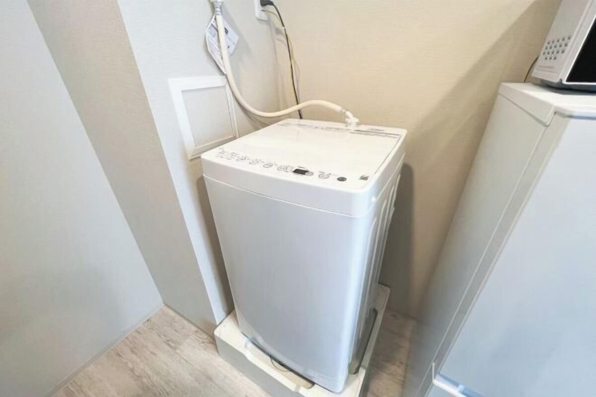 A compact, white, wall-mounted appliance with a control panel, likely a water heater or boiler, in a corner of a room with light-colored walls and a wooden floor.