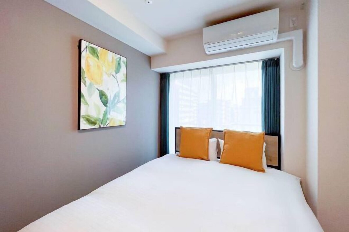 A hotel room with a large bed, two pillows, a window with curtains, an air conditioning unit, and a painting on the wall.