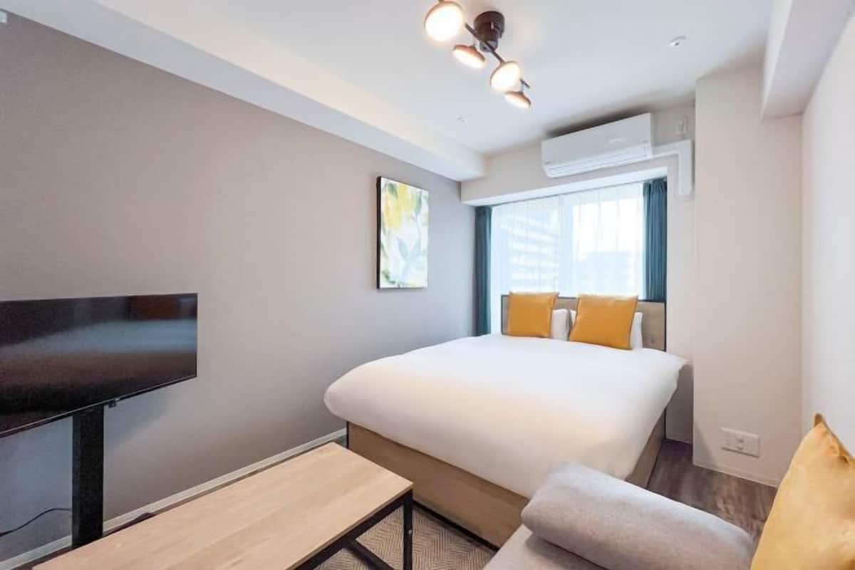 Una habitación de hotel moderna con una cama grande, un televisor de pantalla plana, una mesita pequeña y una ventana con cortinas.