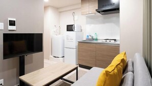 A compact living space with a flat-screen TV, a wooden table, a sofa with a yellow pillow, a kitchenette with a microwave, a refrigerator, and a stove. - LAFESTA Higashi Komagata　 (Tokyo)