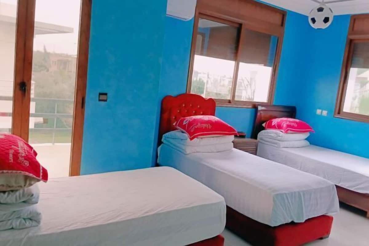 A room with two single beds, one with a red headboard and pillows, and another with a white headboard. There is a window with a view of the outside, and a ceiling fan with a soccer ball decoration.