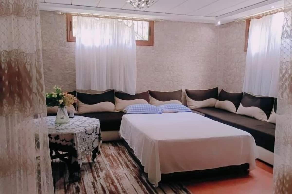 A bedroom with a bed, a small table with a vase, and a window with curtains.