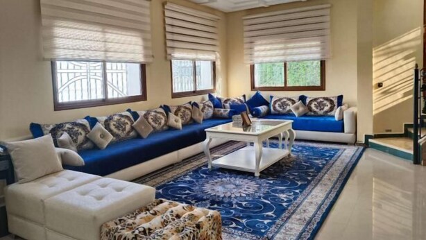 A living room with a blue sofa, white armchair, and patterned ottoman. There is a coffee table and a large window with blinds. - Morocco Casablanca Polar Bear Airport Homestay (Nouaceur)