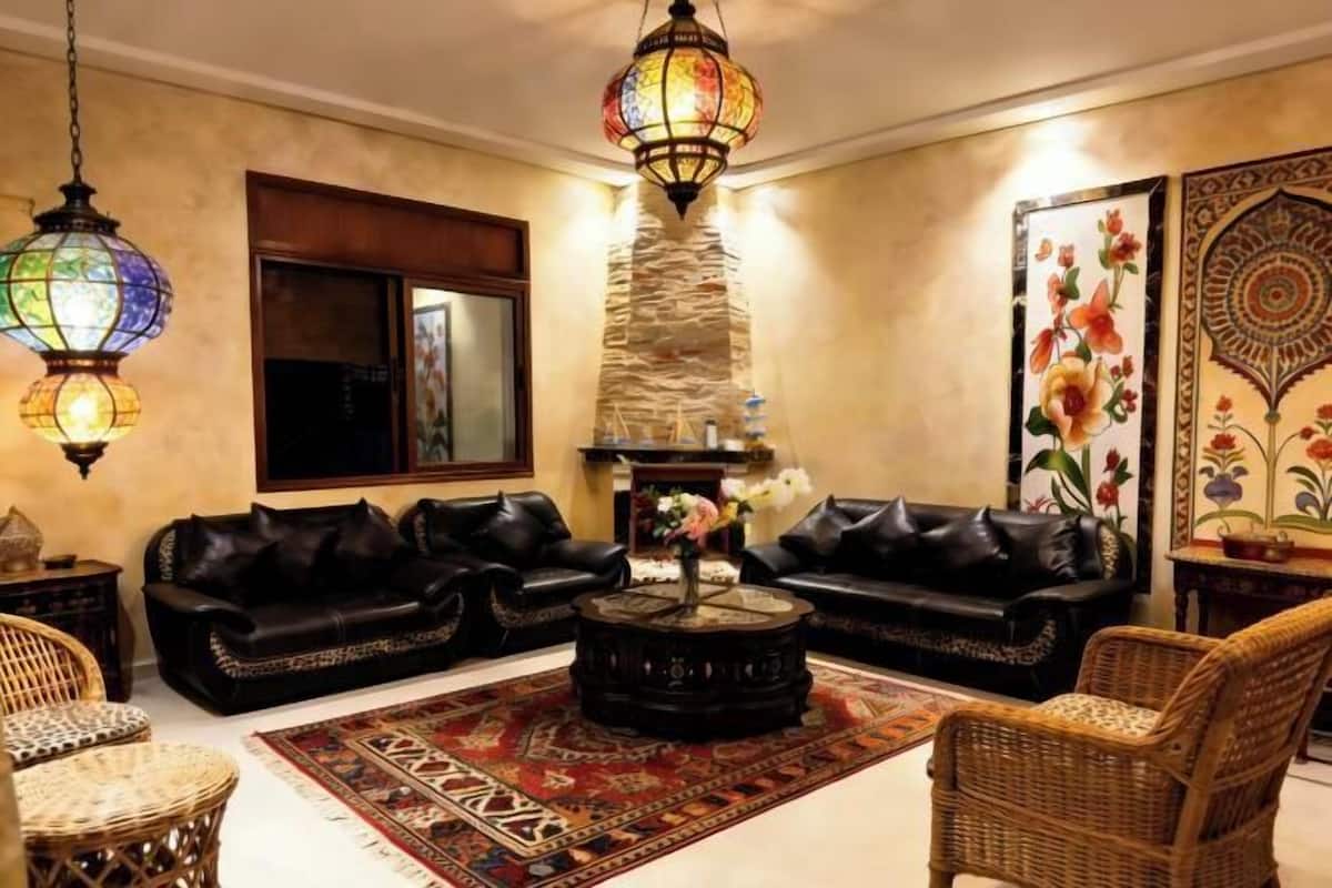 A living room with two black leather sofas, a round coffee table, wicker chairs, a patterned rug, and a decorative wall piece.