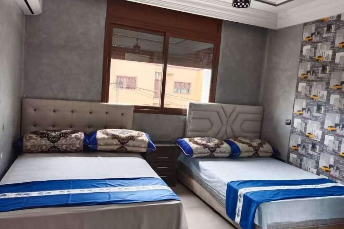 A hotel room with two beds, a window with a view of buildings, and a patterned wall.