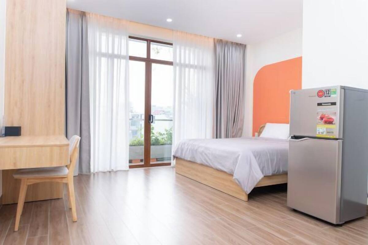 A hotel room with a bed, a desk, a chair, a refrigerator, and a sliding glass door to the outside.