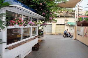 Un puesto de comida en la calle con cestas de flores colgantes, una motocicleta pasando y un edificio al fondo.