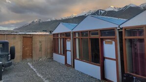 A row of small, single-story buildings with large windows, set against a backdrop of snow-capped mountains under a partly cloudy sky. - Alpine Cottage Pangong (Spangmik)