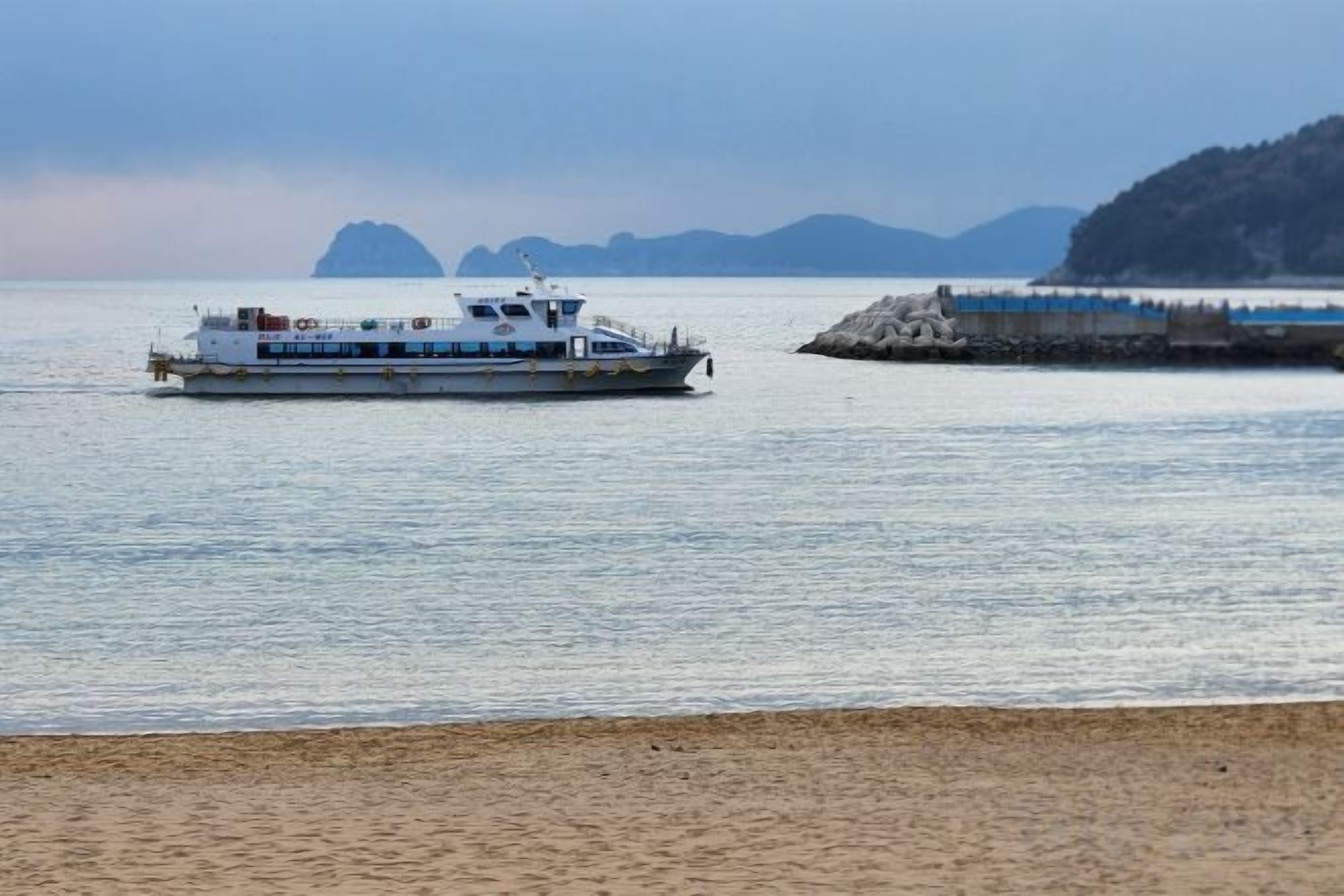 A ferry boat on calm water with a beach and distant mountains in the background.