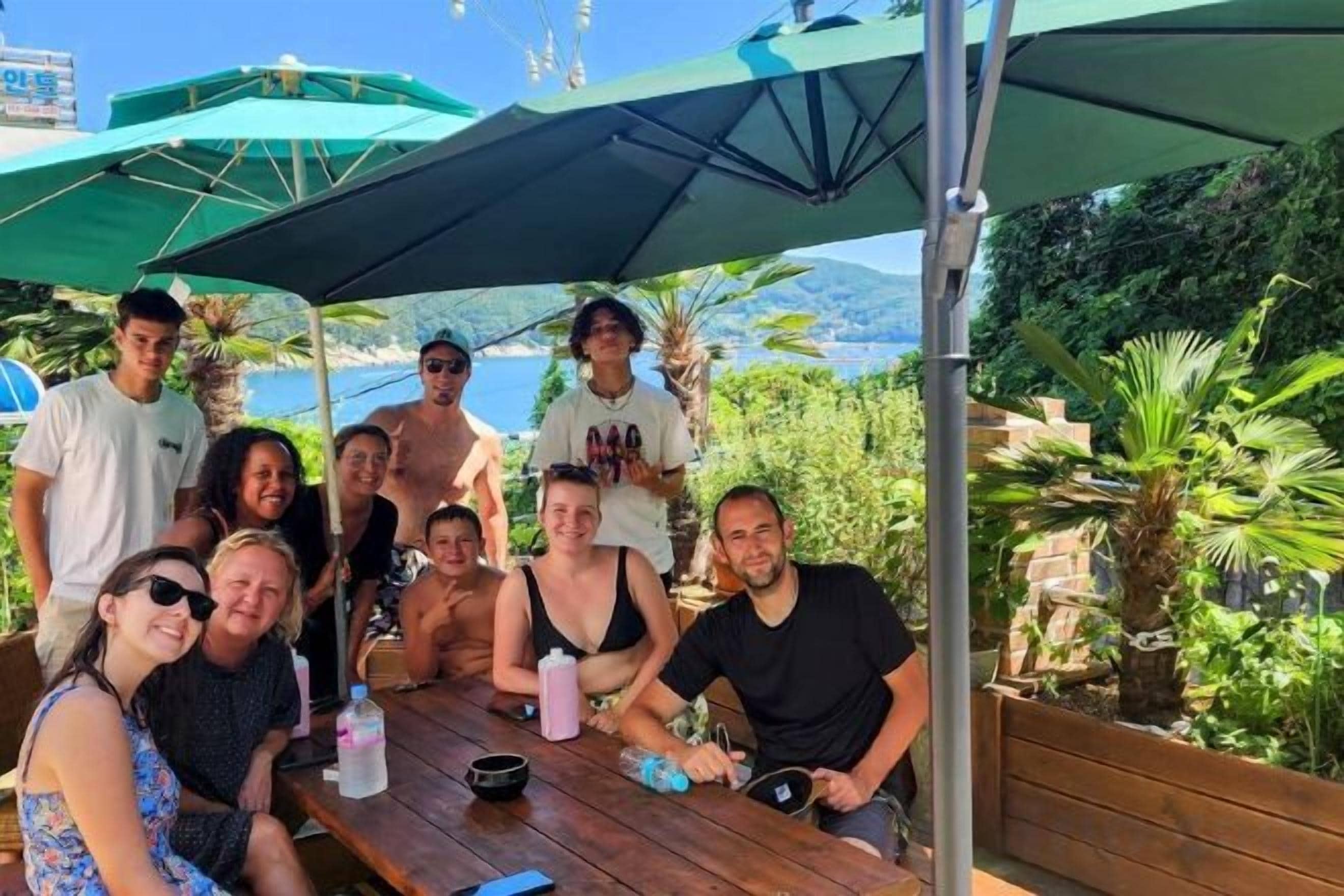 Een groep mensen zit aan een buitentafel met parasols, omgeven door groen en een uitzicht op zee.