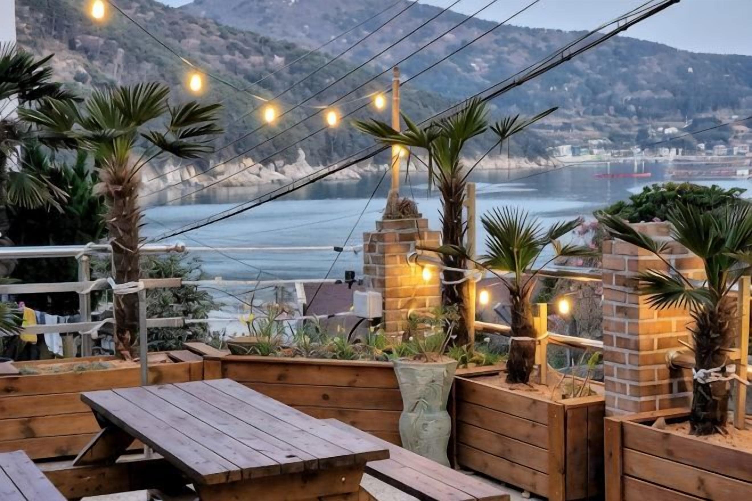 An outdoor seating area with wooden tables and benches, surrounded by plants and brick structures, overlooking a coastal landscape.