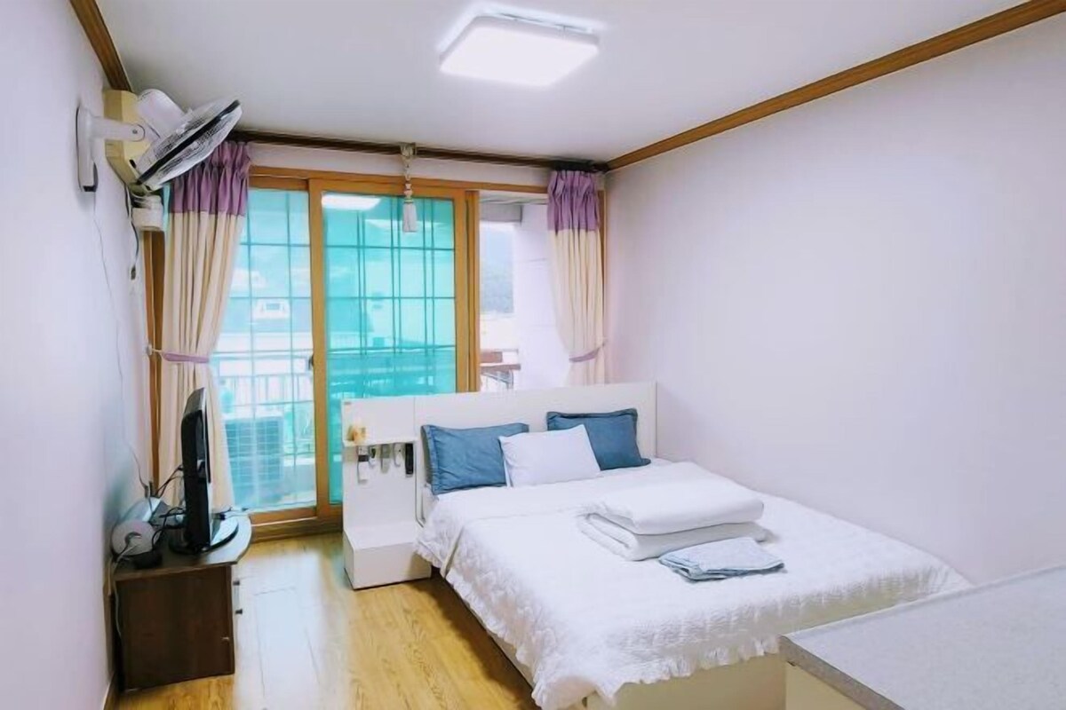 A bedroom with a bed, a desk with a computer, a window with curtains, and a ceiling fan.