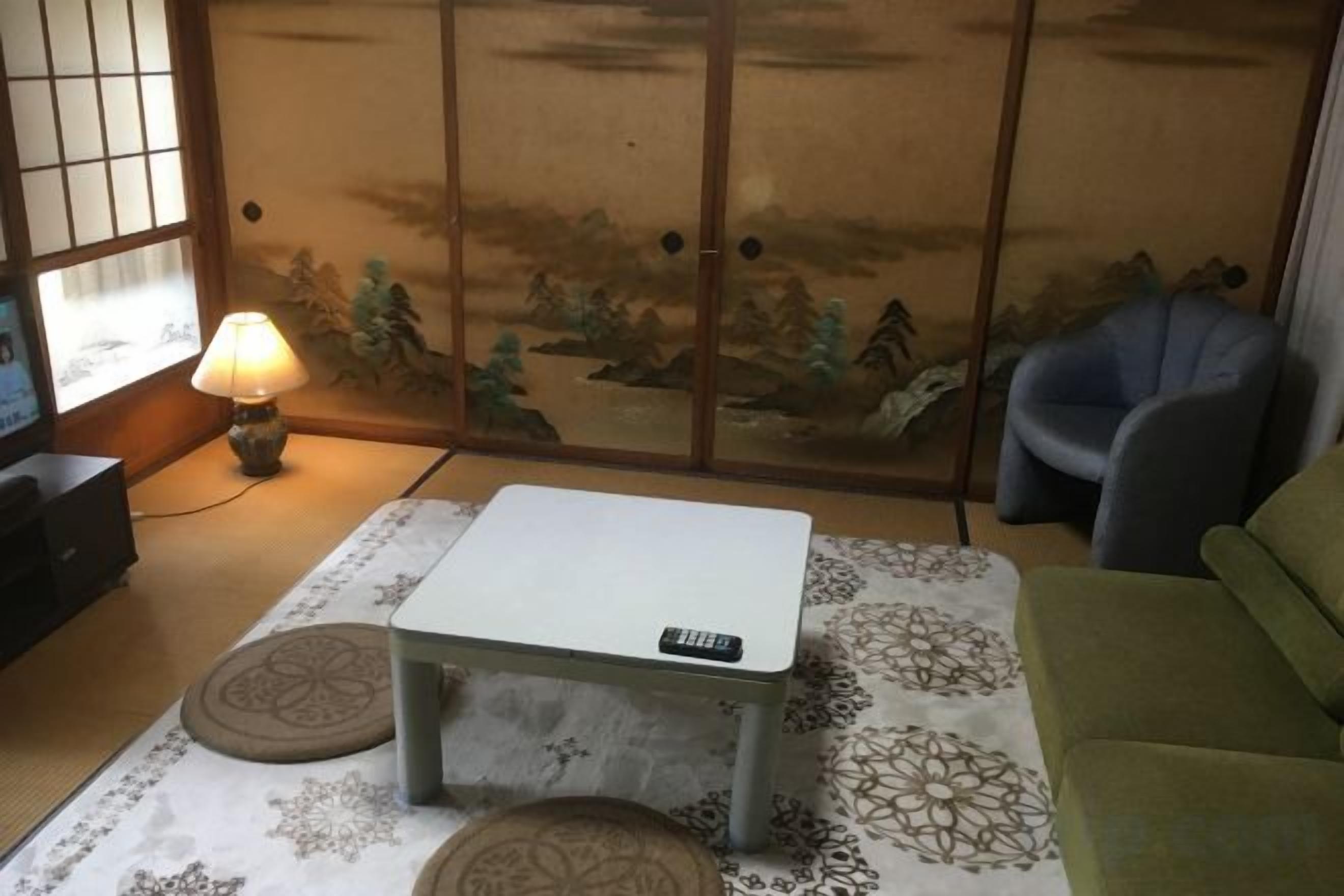Quarto com piso de tatame, esteira de tatame no centro, mesa baixa com controle remoto, sofá verde e porta tradicional deslizante com pintura de paisagem.