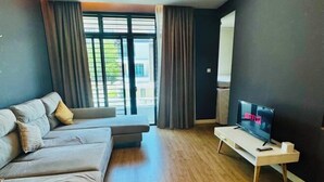 Een moderne woonkamer met een bank, een televisie op een dressoir en grote ramen met gordijnen.