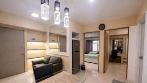 A modern living room with a glass dining table, a sofa, a refrigerator, and a clock on the wall. - Sweet Home (2 Bedroom Apartment) (Mulyorejo)