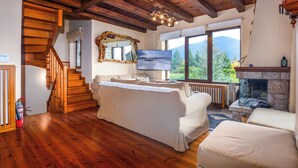 Un soggiorno con pavimento in legno, un divano, un caminetto, una televisione e un'ampia finestra che si affaccia su un paesaggio montuoso.