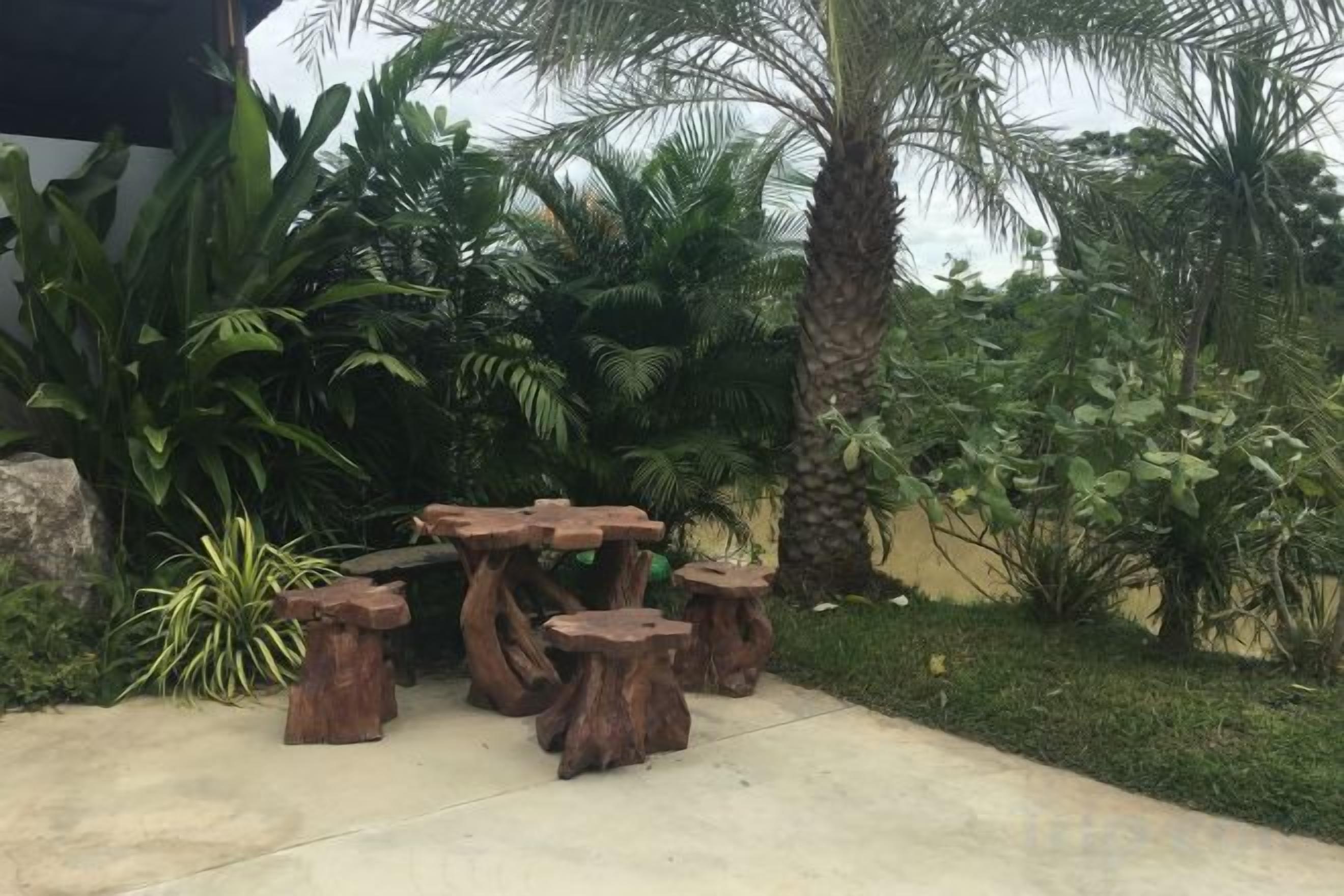 A concrete patio with a rustic wooden table and stools, surrounded by lush tropical plants.