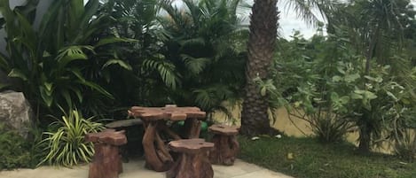 A concrete patio with a rustic wooden table and stools, surrounded by lush tropical plants.