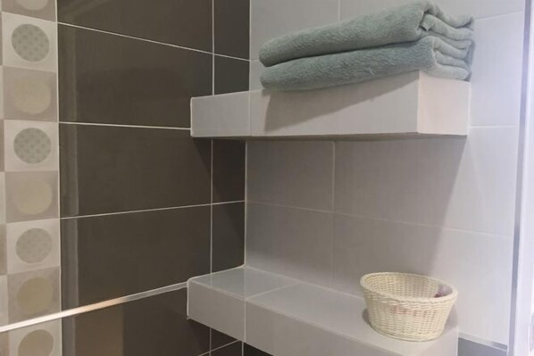 Un bagno con piastrelle scure, uno scaffale bianco con un asciugamano piegato e un cesto in vimini.