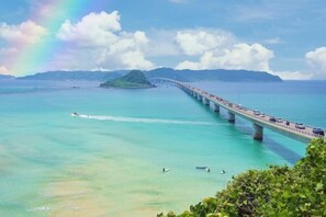 Een lange brug over turquoise water, met een regenboog, een bootje en een klein eiland in de verte.