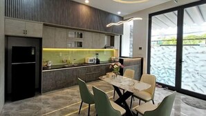 A modern kitchen with a dining area, featuring a marble table and chairs, a built-in oven, and a large window with a view of greenery. - Aroma Beach (Da Nang)
