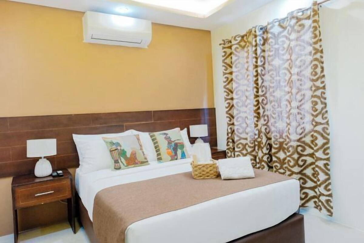 Une chambre d’hôtel équipée d’un lit, d’une table de chevet, d’une lampe, d’un climatiseur et de rideaux à motifs.