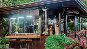 A rustic outdoor bar with a wooden counter and stools, surrounded by greenery.