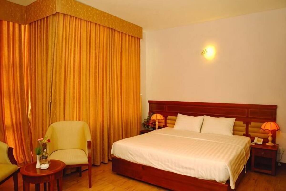 A hotel room with a large bed, a chair, a small table, and a nightstand with a lamp.