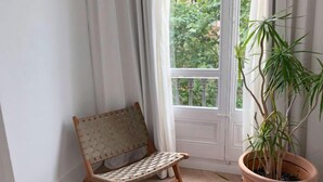 Un coin aménagé avec un fauteuil en osier, une plante en pot et une fenêtre donnant sur un paysage verdoyant.