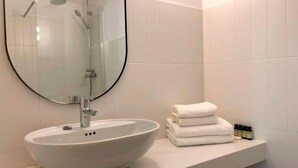 Une salle de bain avec un lavabo blanc, une douche en verre et un miroir. On y trouve des serviettes pliées et des articles de toilette sur le comptoir.