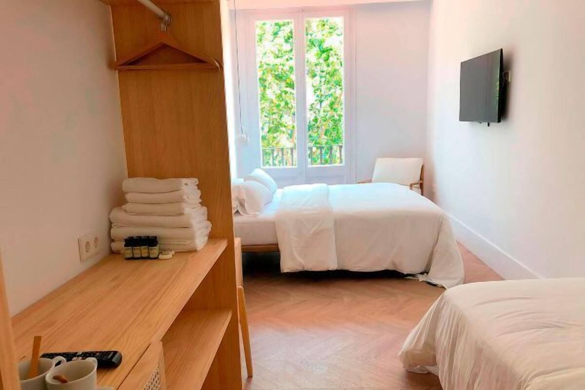 A bedroom with a bed, a wooden shelf with towels, a TV, and a window with a view of greenery.