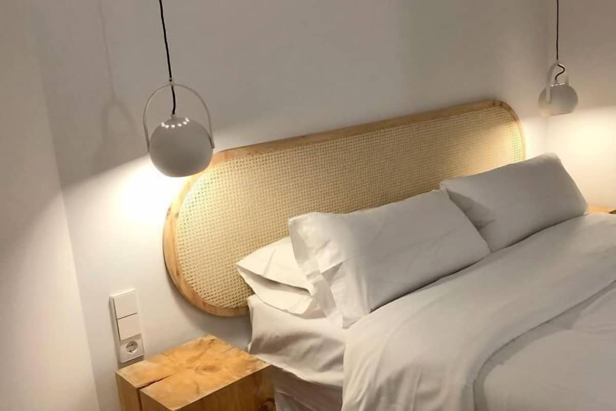 A neatly made bed with white linens, a wooden headboard, and two pendant lights.
